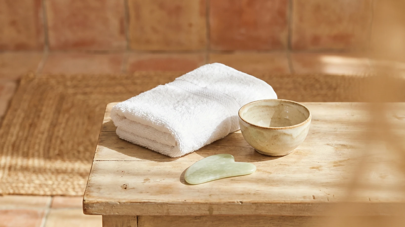 Tovallola blanca neta, un bol de ceràmica i una eina facial de pedra llisa sense marca sobre una superfície clara, amb llum natural suau